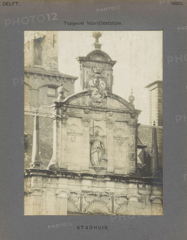 Town Hall. Gable Front (East) Side, Gable of the town hall in Delft with a sculpture of Lady Justice and a coat of arms with a lion, photograph, inscription: 'Delft. 1620.', number: '51', inscription: 'The right of reproduction of these photos is reserved by the Publisher.', Delft, c. 1900 - c. 1904, photographer, paper, pasteboard, gelatin silver print, height 157 mm x width 118 mm, town hall, coat of arms (as symbol of the state, etc.), balustrade, 'Giudice' (Ripa), A sepia-toned photograph of an ornate stone building facade framed by a grey mat, showing carved architectural details, rounded arches, decorative scrollwork, and sculpted stone figures. The upper portion features a central decorative crest with a sculpted animal relief topped by a rounded ornament and flanked by finials. Below the crest is a recessed niche containing a standing statue draped in flowing garments, holding scales. The facade shows pilasters, carved capitals, and shell-shaped motifs above arched panels, with...