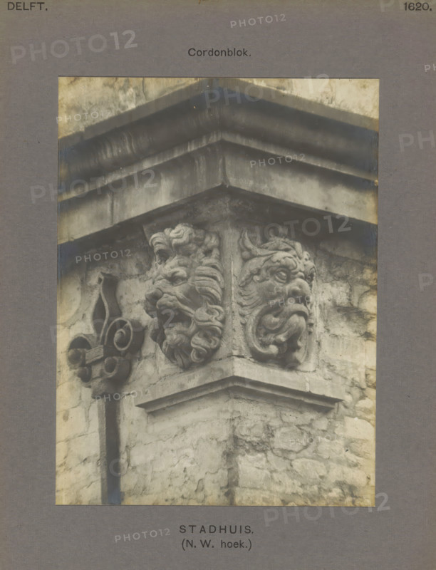 Town Hall. (NW corner.) Cordon block, Cornerstones of the town hall in Delft, decorated with a lion's head and a fantasy head, photograph, inscription: 'Delft. 1620.', number: '54', inscription: 'The right of reproduction of these photos is reserved by the Publisher.', Delft, c. 1900 - c. 1904, photographer, paper, pasteboard, gelatin silver print, height 155 mm x width 116 mm, townhall, lion's head, ornament, hybrid animals, Stadhuis (Delft), A close view of a stone corner of a building showing a carved architectural block with two sculpted grotesque faces and ornate scrollwork, set beneath layered projecting cornices; the stone surface shows weathering and discoloration with visible mortar lines and textured masonry.