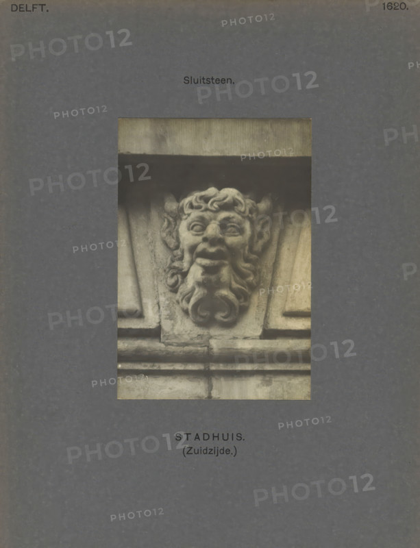Town Hall (South side.) Keystone, Keystone of the town hall in Delft, decorated with a head of presumably a faun, photograph, inscription: 'The right of reproduction of these photos is reserved by the Publisher.', inscription: 'Delft. 1620', number: '57', Delft, c. 1900 - c. 1904, photographer, paper, pasteboard, gelatin silver print, height 108 mm x width 75 mm, townhall, satyrs, fauns, Sileni, keystone, architecture, Town Hall (Delft), A central rectangular photograph shows a carved stone grotesque with a stylized face featuring curled hair, prominent ears, wide eyes, an open mouth, and a curled beard set within architectural stonework; the photograph is mounted on a dark textured background with small printed labels above and below the image.