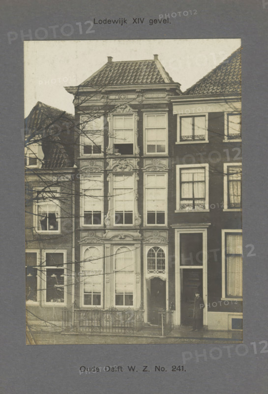 View of a building on the Oude Delft in Delft, Oude Delft WZ No. 241. Louis XIV facade, photograph, inscription: 'The right of reproduction of these photos is reserved by the Publisher.', inscription: 'Delft. 1741.', number: '62', Delft, c. 1900 - c. 1904, photographer, paper, pasteboard, gelatin silver print, developed gelatin silver print, height 154 mm x width 110 mm, urban housing, Oude Delft, A sepia photograph of a narrow multi-story building with ornate carved stone window surrounds and decorative panels, flanked by adjacent brick buildings with tiled roofs; the central building has tall sash windows with shutters and a rounded arched doorway with intricate glazing above it, a small fenced area in front, bare tree branches crossing the façade, and a man standing at the entrance of the neighboring building wearing a dark overcoat and hat.