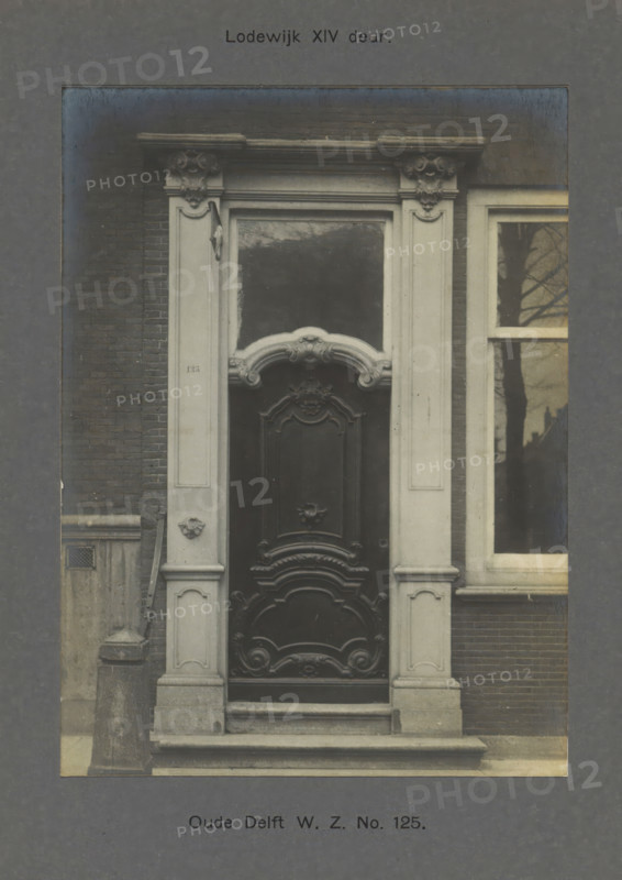 Oude Delft WZ No. 125. Louis XIV door, Door of a building on the Oude Delft in Delft, photograph, inscription: 'Delft. 18th century.', number: '63', inscription: 'The right of reproduction of these photos is reserved by the Publisher.', Delft, c. 1900 - c. 1904, photographer, paper, pasteboard, gelatin silver print, developed gelatin silver print, height 156 mm x width 115 mm, urban housing, door, Oude Delft, A centered ornate wooden door with carved panels and decorative scrollwork set within a pale stone surround featuring fluted pilasters and sculpted capitals, a curved ornamental pediment above a rectangular transom, stone steps leading up to the threshold, a brick wall flanking the doorway on the left, a tall window with a reflection of bare tree branches to the right, and a stone bollard and small vent visible near the base on the left.