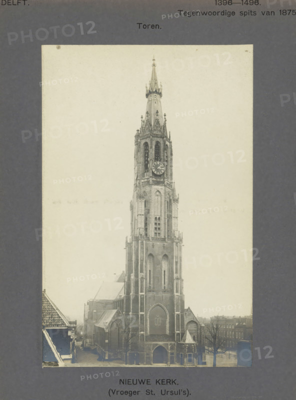 View of the Nieuwe Kerk in Delft, Nieuwe Kerk. (Formerly St. Ursul's). Tower, photograph, inscription: 'The right of reproduction of these photos is reserved by the Publisher.', number: '7', inscription: 'Delft. 1396-1496. Present spire of 1875.', Delft, c. 1900 - c. 1904, photographer, paper, pasteboard, gelatin silver print, developed gelatin silver print, height 166 mm x width 108 mm, church (exterior), parts of church exterior and annexes: tower, Nieuwe Kerk (Delft), A tall Gothic church tower with pointed spire and ornamental pinnacles rises from a large stone church building, showing arched windows, buttresses, a clock face on the tower, and an arched main entrance; surrounding the church are rooftops of nearby buildings, bare trees, and a street visible at the base, all presented within a pale rectangular photograph mounted on a dark textured background.