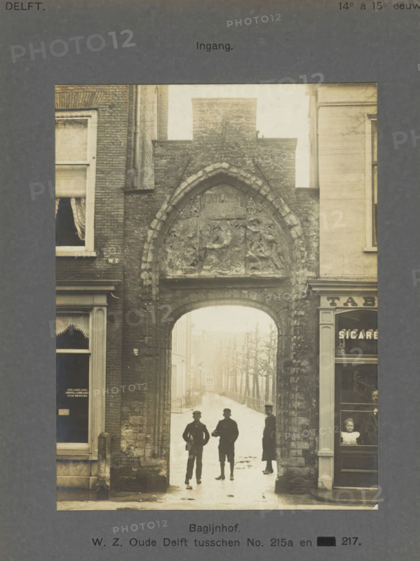 Entrance gate to the Bagijnhof in Delft with three people, Bagijnhof. WZ Oude Delft between No. 215a and 217. Entrance, photograph, inscription: 'Delft. 14th to 15th century.', inscription: 'The right of reproduction of these photos is reserved by the Publisher.', number: '8', Delft, c. 1900 - c. 1904, photographer, paper, pasteboard, gelatin silver print, height 155 mm x width 117 mm, alms-house (Dutch: 'hofje'), gate, entrance, Bagijnhof (Delft), A narrow stone archway set into a brick and masonry street facade frames a view down a tree-lined street; the arch has a pointed top with carved relief panels above and textured stonework, flanked by adjacent building fronts with windows and shopfront details, and three men stand silhouetted beneath the arch while a child is visible through a shop window at the right.