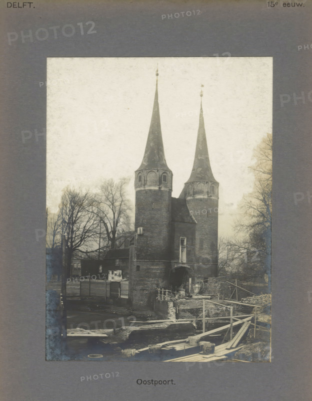 View of the East Gate in Delft, East Gate, photograph, inscription: 'Delft. 15th century.', inscription: 'The right of reproduction of these photos is reserved by the Publisher.', number: '9', Delft, c. 1900 - c. 1904, photographer, paper, pasteboard, gelatin silver print, developed gelatin silver print, height 157 mm x width 116 mm, city-gate, A framed photograph shows a brick gatehouse with two tall conical spires topped with small finials, flanked by bare trees. The gatehouse has rounded towers with small arched windows and a central arched passage at ground level. In front of the structure there is a water channel and a wooden construction of planks, beams, and temporary railings leading toward the foreground. Scattered construction materials and stone blocks are visible near the water, and portions of fencing and a small building appear beside the gatehouse. The sky is pale and overcast, and the overall image has a faded, aged appearance.
