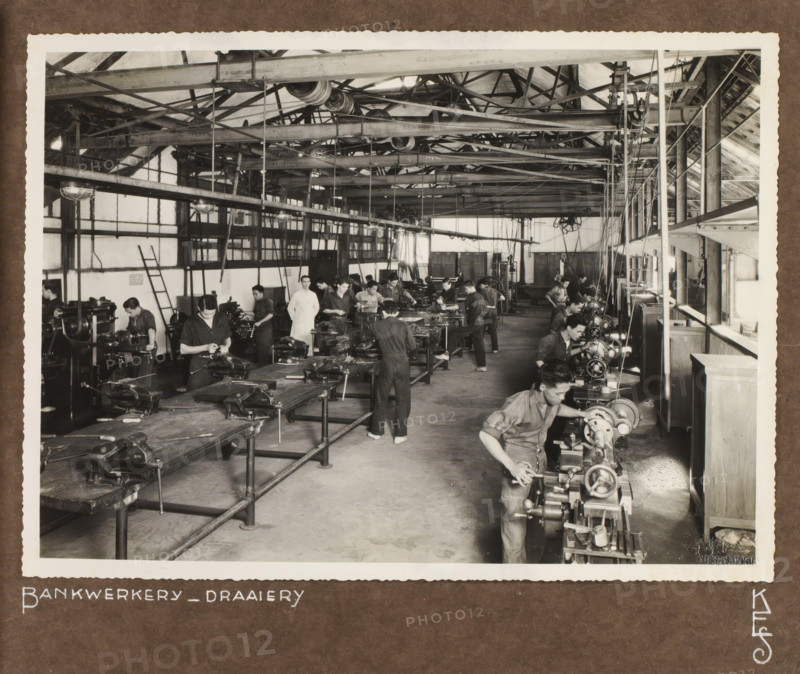Bench workshop in the Queen Emma School, photograph, View of the bench workshop or turning shop in the Queen Emma School in Surabaya where students are working under the supervision of a teacher. Photo pasted into the photo album presented to JHA van der Hoeven upon the completion of his service in Industrial Education in the Dutch East Indies on July 31, 1933., inscription: 'Bankwerkery of Draaiery, KES', photographer: Fotax (Soerabaja), Surabaya, 1933, Java, East Java, paper, gelatin silver print, height 180 mm x width 246 mm, Queen Emma School, founded in 1912, replacing the Civic Evening School that had existed since 1879., Person, JHA van der Hoeven, vocational education, eg: junior technical school, colonial government, cultural history, education, Dutch East Indies, The, Period, 1911 - 1933, A large workshop interior with long wooden workbenches and rows of metal machines, overhead beams and pulleys spanning the ceiling, and multiple men standing or working at the machines weari...