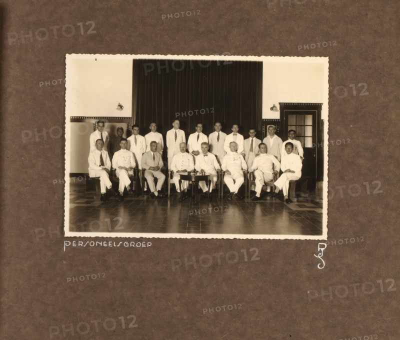 Staff of the Princess Juliana School, photograph, Group photo of the staff of the Princess Juliana School in Yogyakarta. Group of men posing in two rows in the school. Photo pasted into the photo album presented to JHA van der Hoeven upon the completion of his service in Industrial Education in the Dutch East Indies on July 31, 1933., inscription: 'Personnel Group, PJS', Yogyakarta, 1933, photographer, Java, Central Java, paper, gelatin silver print, height 121 mm x width 175 mm, Princess Juliana School in Yogyakarta, founded in 1917, initially intended only as a training program for construction engineers, Person, JHA van der Hoeven, vocational education, eg: junior technical school, anonymous historical persons portrayed in a group, in a group-portrait, colonial government, cultural history, education, Dutch East Indies, The, Period, 1911 - 1933, A sepia photograph shows a group of men posed indoors in front of a dark curtain and light walls with wall-mounted lamps and a doorway with...