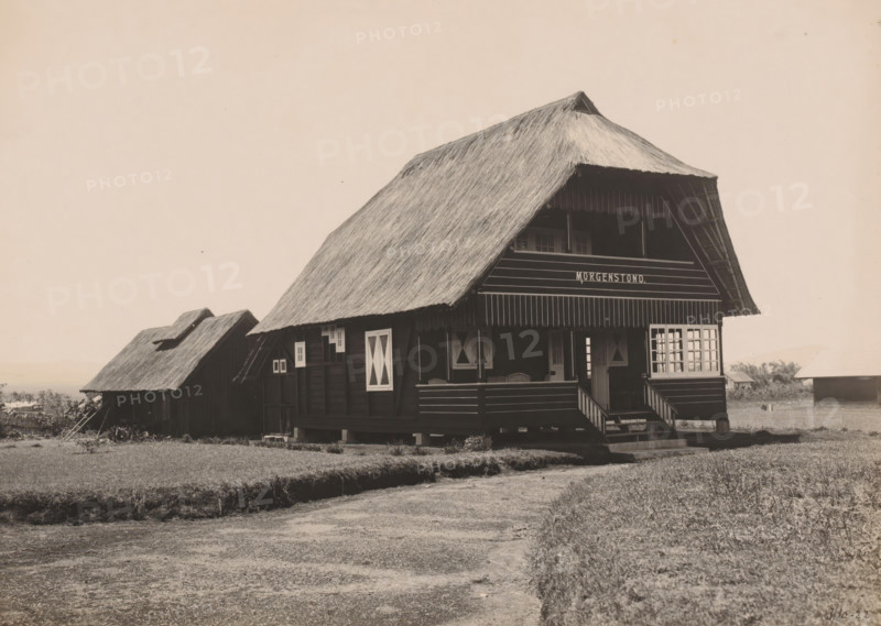 Country house Kaban Djahe, Belawan Ocean Harbor, photograph, View of the country house 'Morgenstond' in Kabandjahe, probably the country house of director Laurens de Groot. Numbered: 230. Part of the group of loose photographs regarding the construction of the port of Belawan on the coast off Medan, Sumatra 1921-1922., date: '3, 10-22', annotation: 'No. 230 Buitenhuis Kaban Djahe', Kabanjahe, 1922-10-03, photographer, Sumatra, paper, gelatin silver print, developing gelatin silver print, height 160 mm x width 220 mm, hut, cabin, lodge, economic history, overseas history, traffic and transport, infrastructure, harbor, dredging, Period, 1921 - 1922, A large wooden house with a steep thatched roof sits on stilts above the ground, featuring a covered porch with steps leading up, shuttered windows with diagonal panels, and a balcony area under the eaves; to the left a smaller thatched structure with a pitched roof is partially visible, and to the right more low buildings can be seen in the ...