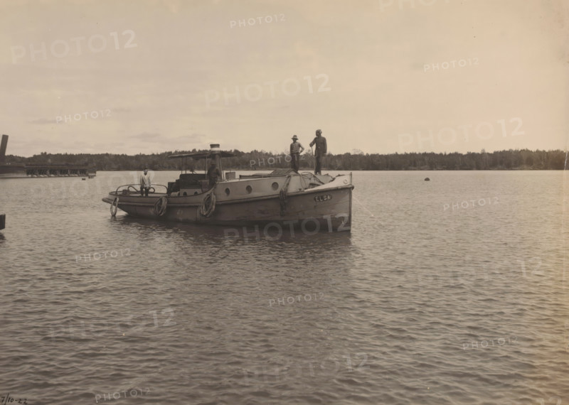 Motor tug 'Elsa', Ocean Harbor Belawan, photograph, The motor tug 'Elsa'. Numbered: 235. Part of the group of individual photographs regarding the construction of the harbor of Belawan on the coast off Medan, Sumatra 1921-1922., annotation: 'No. 235 Motor tug 'Elsa'', date: '7, 10-22', Belawan, 1922-10-07, photographer, Sumatra, paper, gelatin silver print, developing gelatin silver print, height 160 mm x width 220 mm, tug, pusher tug, economic history, overseas history, traffic and transport, infrastructure, harbor, dredging, Period, 1921 - 1922, A small boat floats on calm water with three people standing on its deck; the vessel has a covered central area with a small funnel and round portholes along the hull, coiled ropes draped over the sides, and a lifebuoy hanging at the stern. The boat is positioned near a low treeline that stretches across the background and a wooden pier is visible to one side. The sky is light and mostly clear with a few thin clouds, and the water surface sho...