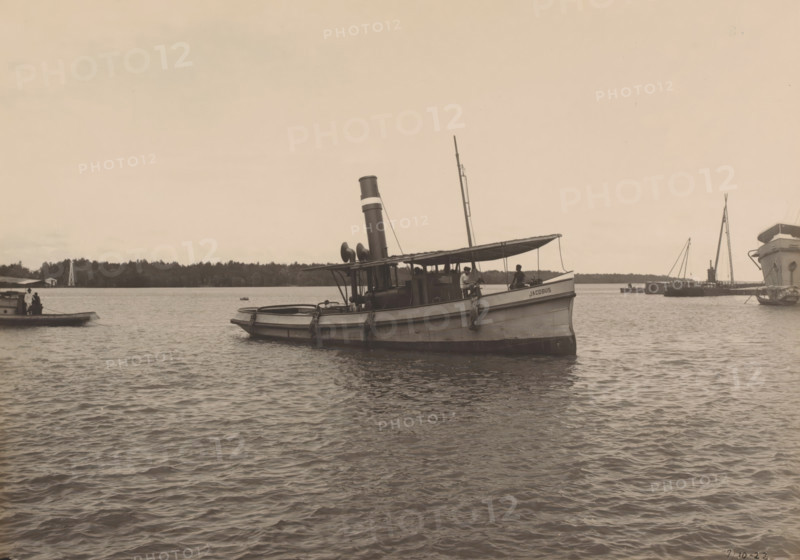 Tugboat 'Jacobus', Ocean Harbor Belawan, photograph, The motor tugboat 'Jacobus'. Numbered: 236. Part of the group of individual photographs regarding the construction of the harbor of Belawan on the coast off Medan, Sumatra 1921-1922., annotation: 'No. 236 Tugboat 'Jacobus'', date: '7, 10-22', Belawan, 1922-10-07, photographer, Sumatra, paper, gelatin silver print, developing gelatin silver print, height 160 mm x width 220 mm, tug, pusher tug, economic history, overseas history, traffic and transport, infrastructure, harbor, dredging, Period, 1921 - 1922, A medium-sized steam launch floats on calm water with a canopy over its midsection, a tall funnel rising behind the canopy, and a mast extending upward toward the sky; two people are visible on the deck, one seated near the bow and another near the cabin, and coiled ropes hang along the hull. In the background a tree-lined shore runs across the horizon with several other boats and masts visible near the right edge of the scene, and a...