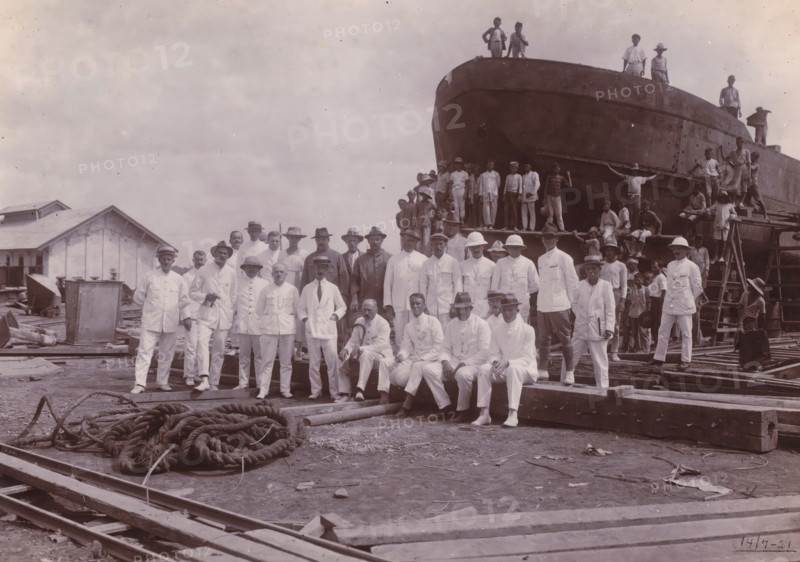 Farewell visit Chief Engineer Fritzlin, Belawan Ocean Port, photograph, Group photo at the construction site in front of the bow of a ship, on the occasion of the farewell visit of Chief Engineer Fritzlin, second director of the port of Belawan, July 14, 1921. In the foreground the Dutch in white uniforms, behind them Indonesian boys on and in front of the ship. Numbered: 47. Part of the group of individual photos regarding the construction of the port of Belawan on the coast off Medan, Sumatra 1921-1922., date: '17, 7-21', inscription: 'No. 47. Ocean Port Belawan, Farewell Visit Chief Engineer Fritzlin', Belawan, 1921-07-14, photographer, Sumatra, paper, daylight gelatin silver print, height 160 mm x width 220 mm, harbor, building-site, economic history, overseas history, traffic and transport, infrastructure, Period, 1921 - 1922, A large group of people pose in front of a partially constructed or repaired large vessel on blocks, with many standing on the ground wearing light-colored ...