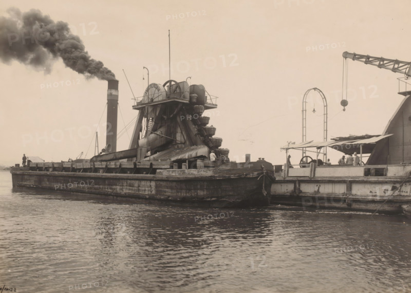 Dredger Amsterdam, Ocean Harbor Belawan, photograph, The dredger 'Amsterdam' on the water next to another ship. Numbered: 240. Part of the group of individual photographs regarding the construction of the harbor of Belawan on the coast off Medan, Sumatra 1921-1922., date: '9, 10-22', inscription: 'No. 240 Baggermolen Amsterdam', Belawan, 1922-10-09, photographer, Sumatra, paper, gelatin silver print, height 160 mm x width 220 mm, building-site, dredge, dredging machine, economic history, overseas history, traffic and transport, infrastructure, harbor, dredging work, Period, 1921 - 1922, A sepia-toned waterfront scene showing a large, flat-topped barge with heavy industrial machinery mounted on it, including a multi-bucket wheel structure with curved chutes and supporting beams, a tall smokestack emitting a dense plume of dark smoke, and various cables and poles rising from the deck. The barge's weathered hull sits low in the water, with visible rails and ladders along its side and a sm...