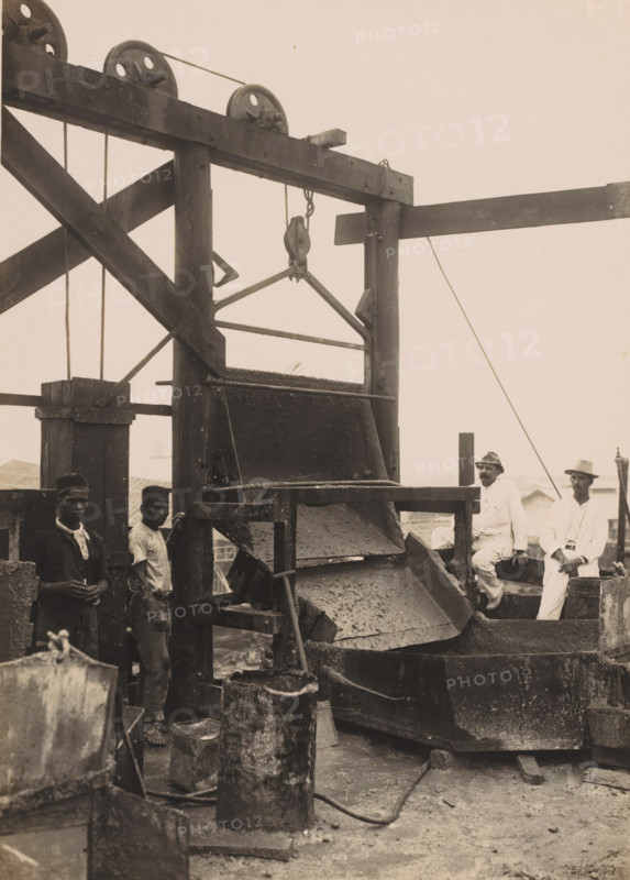 On top of the pouring jetty, Ocean Harbor Belawan, photograph, Scaffolding used to pour the concrete, with workers and supervisors. Numbered: 269. Part of the group of individual photographs regarding the construction of the port of Belawan on the coast off Medan, Sumatra 1921-1922., annotation: 'No. 269 On top of the dumping pier', date: '25, 10-22', Belawan, 1922-10-25, photographer, Sumatra, paper, gelatin silver print, gelatin silver developing print, height 220 mm x width 160 mm, building-site, making the overground structures (, building activities), economic history, overseas history, traffic and transport, infrastructure, harbor, dredging, Period, 1921 - 1922, A large wooden industrial structure with pulleys and ropes dominates the scene, showing a chute or hopper with layers of rough material cascading into a basin below, with wooden beams and diagonal braces forming a frame around it; several metal buckets and cylindrical containers sit on the ground in front of the machinery...