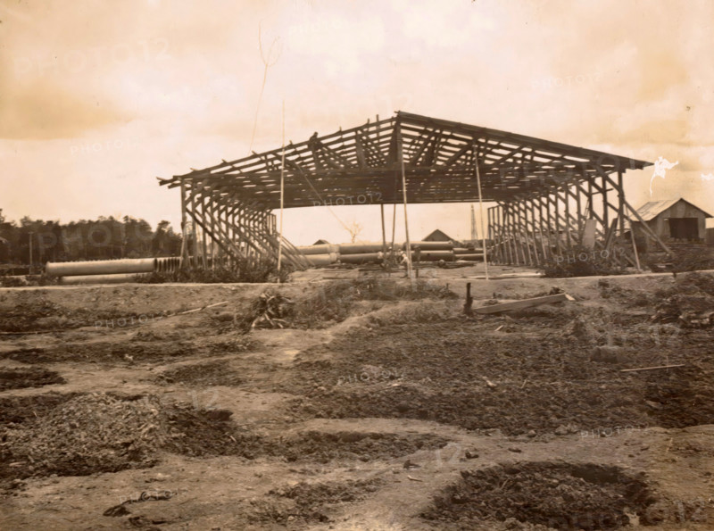 Construction of a shed, photograph, Framework of a shed under construction. Part of the group of individual photographs regarding the construction of the port of Belawan on the coast off Medan, Sumatra 1921-1922., Belawan, 1921 - 1922, photographer, Sumatra, paper, gelatin silver print, height 125 mm x width 170 mm, building-site, making the overground structures (, building activities), economic history, overseas history, traffic and transport, infrastructure, harbor, dredging, Period, A large wooden framework of a structure stands centrally with exposed rafters and supporting posts, flanked by vertical braces on both sides, and a smaller building with a pitched roof is visible to the right; cylindrical pipes lie on the ground beneath the framework and extend toward the foreground, while the ground in front appears uneven with disturbed soil and scattered debris, and a treeline runs across the background beneath a pale sky.