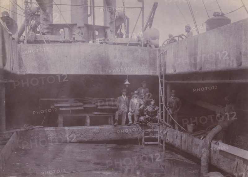 Japanese divers on hold II, photograph, A Japanese diving team poses at the edge of the hold of a dredger or suction vessel. Part of the group of individual photographs regarding the construction of the port of Belawan on the coast off Medan, Sumatra 1921-1922., inscription: 'Japanese divers on hold II', Belawan, 1921 - 1922, photographer, Sumatra, paper, daylight gelatin silver print, height 144 mm x width 198 mm, dredge, dredging machine, Japanese, deep-sea diving, economic history, overseas history, traffic and transport, infrastructure, harbor, dredging, Period, A dimly lit ship interior with a large open hold and concrete-like walls, showing a group of workers standing and sitting on wooden planks and ladders; heavy machinery, winches, ropes, pipes, and a flexible hose run along the edges, and a diving helmet and associated gear rest near the ladder; a hanging lamp is suspended above the group, and additional crew members are visible on the upper deck beside large cylindrical equi...