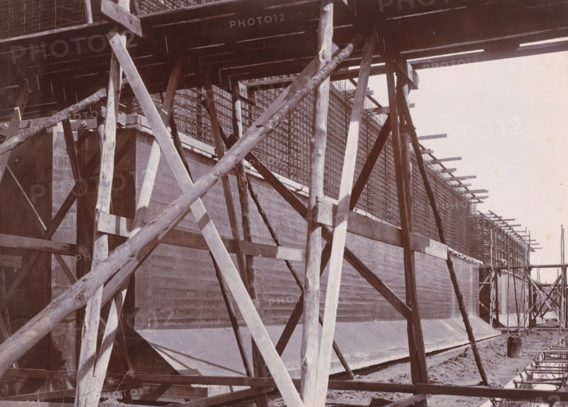 View of caissons 1-5 & 9, Belawan Ocean Harbor, photograph, Construction of the caissons for the construction of the harbor. Numbered: 64. Part of the group of loose photographs of the construction of Belawan Harbor on the coast off Medan, Sumatra 1921-1922., inscription: 'No. 64, Ocean Harbor Belawan, View of caisson 1-5 & 9', date: '3, 10 21', Belawan, 1921-10-03, photographer, Sumatra, paper, daylight gelatin silver print, height 160 mm x width 220 mm, harbor, building-site, economic history, overseas history, traffic and transport, infrastructure, Period, 1921 - 1922, A large wooden scaffold structure with diagonal and vertical beams is visible in front of a tall concrete wall; timber planks form platforms and supports, and rough logs are used as braces. Wire mesh reinforcement sits atop the concrete wall, extending horizontally along its length. Shadows cast by the beams fall across the wall and platforms. Dirt and scattered construction debris lie at the base of the wall, and add...