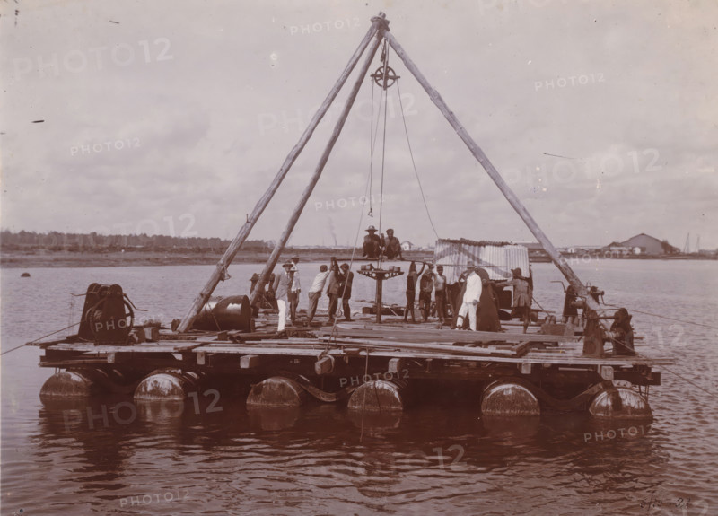 Boiling rig in the channel, Belawan Ocean Harbor, photograph, Floating raft with a drilling rig, at work in the channel of the new harbor, October 1921. Numbered: 67. Part of the group of loose photographs regarding the construction of the port of Belawan on the coast off Medan, Sumatra 1921-1922., inscription: 'No 67 Belawan Ocean Harbor, Boiling rig in the channel', date: '5, 10-21', Belawan, 1921-10-05, photographer, Sumatra, paper, daylight gelatin silver print, height 160 mm x width 220 mm, harbor, building-site, economic history, overseas history, traffic and transport, infrastructure, Period, 1921 - 1922, A large wooden raft floats on calm water with cylindrical flotation drums visible along its underside and simple mechanical winches mounted near its corners, a tall tripod of rough wooden poles rises from the raft center supporting a pulley and hanging cables, and several men in varied clothing stand and sit on the raft around a suspended platform while one man in light clothin...