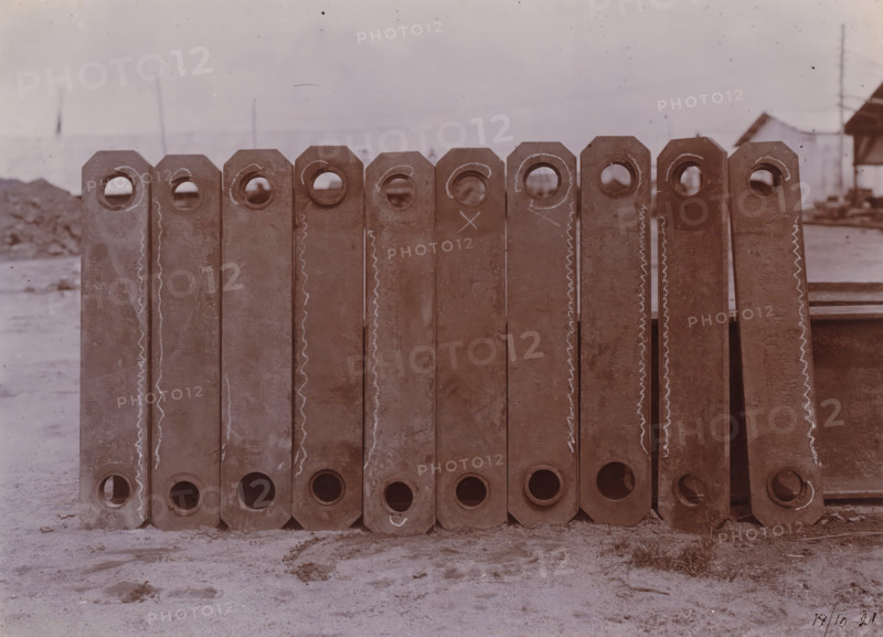 Bottle chain links, Belawan Ocean Harbor, photograph, Parts of the bucket chain of a dredging ship. Numbered: 70. Part of the group of loose photographs about the construction of the port of Belawan on the coast off Medan, Sumatra 1921-1922., inscription: 'No. 70, Bucket chain links', date: '17, 10-21', Belawan, 1921-10-17, photographer, Sumatra, paper, daylight gelatin silver print, height 160 mm x width 220 mm, harbor, building-site, economic history, overseas history, traffic and transport, infrastructure, Period, 1921 - 1922, A row of vertically oriented rectangular metal beams with octagonal tops and circular holes near the top and bottom edges, standing side by side on a sandy surface; the beams show worn and rusty surfaces with chalk-like wavy lines and circular marks on some faces; faint background elements include a distant low horizon, indistinct structures, and utility poles under a pale sky.