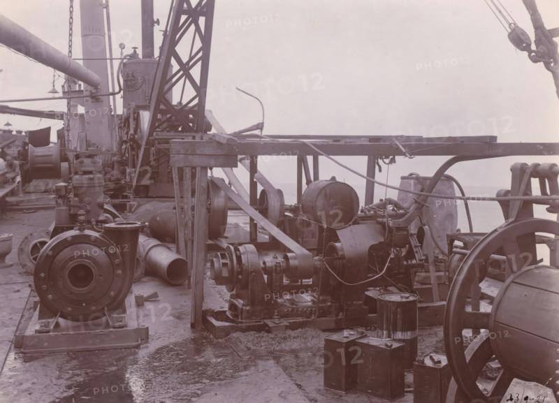 Image of SS Altenburg at Singapore, Belawan Ocean Harbor, photograph, Machinery on deck of the SS Altenburg at Singapore. Numbered: 76. Part of the group of individual photographs regarding the construction of the port of Belawan on the coast off Medan, Sumatra 1921-1922., date: '23, 9-21', inscription: 'No. 76, Image of SS Altenburg at Singapore', Singapore, 1921-09-23, photographer, paper, daylight gelatin silver print, height 160 mm x width 220 mm, steamsailer, economic history, overseas history, traffic and transport, infrastructure, harbor, Sumatra, Belawan, Period, 1921 - 1922, Machinery and heavy metal components fill the deck, including large cylindrical and drum-shaped pieces, pulleys, belts, and pipes arranged among wooden beams and metal supports, with ropes and chains draped across the structure; a lattice metal crane or derrick rises near the center, flanked by vertical columns and mounted equipment, while various fittings, flanges, and housings rest on the deck surface th...