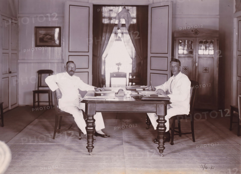 Directors of Grotius Company, Belawan Ocean Port, photograph, Interior with the two directors of the Grotius Contracting Company, sitting behind a work table. Left Laurens de Groot, right probably PJF van Buuren, October 1921. Numbered: 82. Part of the group of loose photographs concerning the construction of the port of Belawan on the coast off Medan, Sumatra 1921-1922., date: '16, 10-21', inscription: 'No 82, Directeuren Mij Grotius', Belawan, 1921-10-16, photographer, Sumatra, paper, daylight gelatin silver print, height 160 mm x width 220 mm, Person, Laurens de Groot, historical persons, office (, administration), director, boss (in modern industry), economic history, overseas history, traffic and transport, infrastructure, harbor, Period, 1921 - 1922, Two men in light-colored suits sit opposite each other at a wooden table with turned legs, papers and writing materials spread across its surface. The man on the left leans back slightly with one arm resting on the chair back and the...