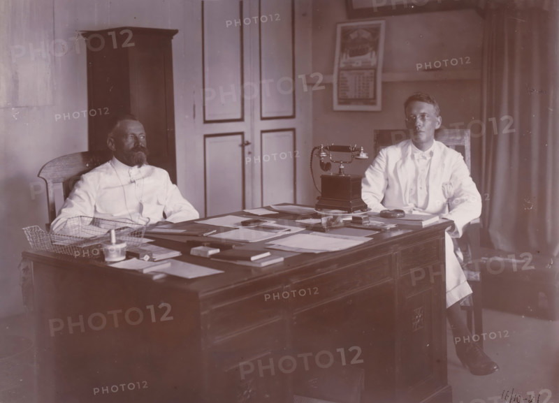 Directors of Grotius Company, Belawan Ocean Port, photograph, Interior with the two directors of the Grotius Contracting Company, sitting behind a desk with a telephone. Left Laurens de Groot, right probably PJF van Buuren, October 1921. Numbered: 83. Part of the group of loose photographs concerning the construction of the port of Belawan on the coast off Medan, Sumatra 1921-1922., date: '16, 10-21', inscription: 'No 83, Directeuren Mij Grotius', Belawan, 1921-10-16, photographer, Sumatra, paper, daylight gelatin silver print, height 160 mm x width 220 mm, Person, Laurens de Groot, historical persons, office (, administration), director, boss (in modern industry), economic history, overseas history, traffic and transport, infrastructure, harbor, Period, 1921 - 1922, An office scene showing two men seated behind a wooden desk covered with scattered papers and stationery, one man with a beard sitting on the left and another clean-shaven man wearing glasses seated on the right, both dres...