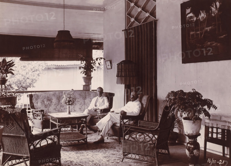 Directors of the Grotius Contracting Company, Oceaanhaven Belawan, photograph, Interior of a house with the two directors of the Grotius Contracting Company, Laurens de Groot on the right, probably PJF van Buuren on the left, October 1921. Untitled and unnumbered. Part of the group of individual photographs concerning the construction of the port of Belawan on the coast off Medan, Sumatra 1921-1922., date: '16, 10-21', Belawan, 1921-10-16, photographer, Sumatra, paper, daylight gelatin silver print, height 160 mm x width 220 mm, Person, Laurens de Groot, office (, administration), director, boss (in modern industry), economic history, overseas history, traffic and transport, infrastructure, harbor, Period, 1921 - 1922, A covered porch or veranda with wicker furniture including chairs, a round table, and a low ottoman, patterned upholstery on a settee, potted plants on stands and on the floor, two men seated in armchairs wearing light-colored clothing, a hanging fringed lampshade and a ...