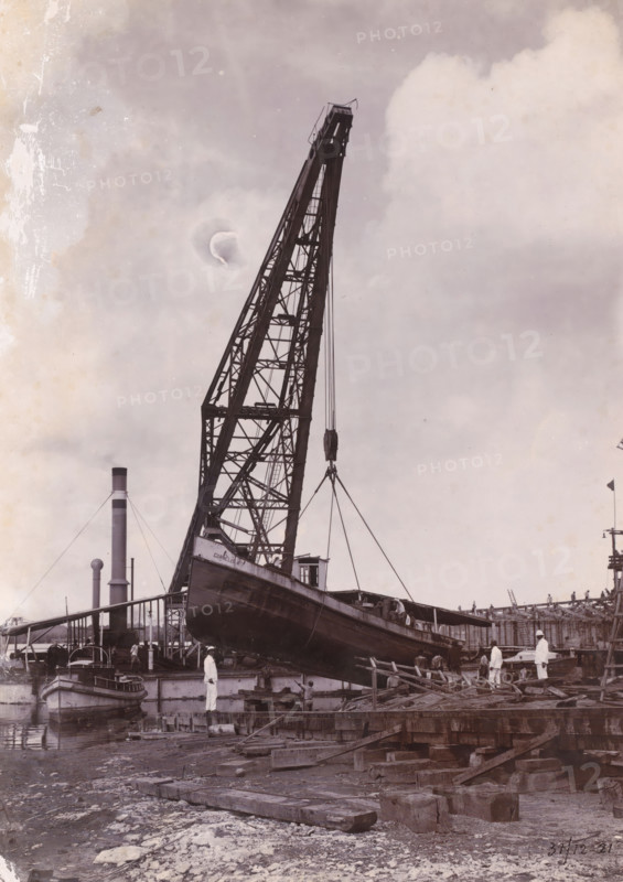 Laying out the tugboat 'Cornelis' to be repaired, Oceaanhaven Belawan, photograph, A tugboat is being lifted out of the water for repair with a crane. Numbered: 105. Part of the group of loose photographs concerning the construction of the port of Belawan on the coast off Medan, Sumatra 1921-1922., inscription: 'No 105 Ocean Port Belawan, Laying down of the tugboat 'Cornelis' to be repaired', date: '31, 12-21', Belawan, 1921-12-31, photographer, Sumatra, paper, daylight gelatin silver print, height 220 mm x width 160 mm, harbour, building-site, dock crane, repairing, caulking, careening (, ship), tug, pusher tug, economic history, overseas history, traffic and transport, infrastructure, harbor, dredging, Period, 1921 - 1922, A large industrial crane with a lattice boom towers upward, its cables suspending a wooden vessel that is angled over a dock; several men in light-colored work clothes stand on the dock and near the hull, and a small boat is moored alongside the platform; in the ba...
