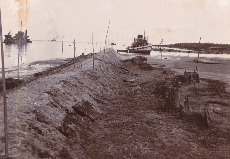 Dumping SS Madjoe (sand loading) 1st work, Belawan Ocean Port, photograph, Dredging and pumping work by the SS Madjoe at the construction pit. Numbered: 133. Part of the group of loose photographs regarding the construction of the port of Belawan on the coast off Medan, Sumatra 1921-1922., inscription: 'No. 133 Ocean Port Belawan, SS Madjoe Dump (sand load) 1st work', date: '11, 3.22', Belawan, 1922-03-11, photographer, Sumatra, paper, daylight gelatin silver print, height 160 mm x width 220 mm, harbor, building-site, dredge, dredging machine, economic history, overseas history, traffic and transport, infrastructure, dredging work, Period, 1921 - 1922, A wide, low-angle shoreline scene showing a raised, narrow earthen embankment running toward the water, with thin vertical poles driven along its length and rough, muddy ground on either side scattered with broken wooden planks and stumps; a large vessel sits moored near the end of the embankment with a smaller boat alongside it, other b...