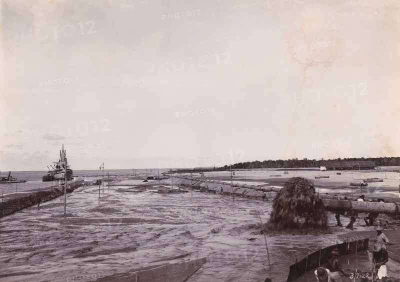 Sand loading by GG Idenburg. Height 2m + BP.), Belawan Ocean Harbour, photograph, Sand loading by the GG Idenburg. Numbered: 176. Part of the group of loose photographs regarding the construction of the port of Belawan on the coast off Medan, Sumatra 1921-1922., date: '3, 7-22', inscription: 'No 176 Sand loading by GG Idenburg. Height 2m + BP.)', Belawan, 1922-07-03, photographer, Sumatra, paper, daylight gelatin silver print, height 160 mm x width 220 mm, harbor, building-site, economic history, overseas history, traffic and transport, infrastructure, dredging, Period, 1921 - 1922, A wide shoreline scene showing wet sand and shallow water with a series of long pipes and wooden supports stretching toward the horizon, a large pile of dark material pouring from the end of a pipe into the foreground, a ship moored near a rocky jetty at the left edge, scattered small boats near the water, several small figures standing or working along the shoreline and on the jetty, low structures and a l...