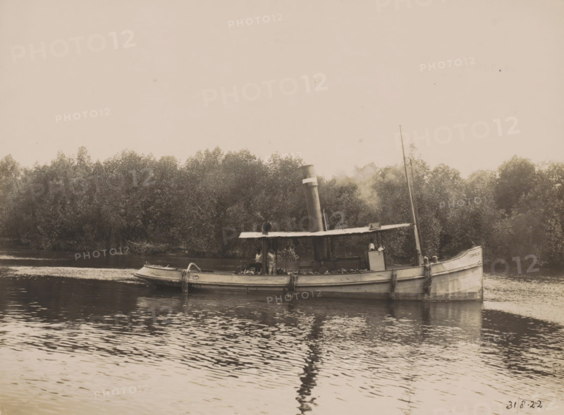 Tugboat 'Cornelis', Ocean Harbor Belawan, photograph, The tugboat 'Cornelis'. Numbered: 192. Part of the group of individual photographs regarding the construction of the harbor of Belawan on the coast off Medan, Sumatra 1921-1922., inscription: 'No. 191, Tugboat 'Cornelis'', date: '31, 8-22', Belawan, 1922-08-31, photographer, Sumatra, paper, gelatin silver print, height 160 mm x width 220 mm, harbor, building-site, tug, pusher tug, economic history, overseas history, traffic and transport, infrastructure, dredging, Period, 1921 - 1922, A weathered steam launch floats on calm water near a tree-lined riverbank, its hull showing signs of age and ropes draped over the side, a covered central cabin topped by a vertical smokestack emitting a faint plume, a small boat tied alongside, a slender mast rising toward the sky, and trees densely filling the background while ripples and reflections ripple across the water surface.