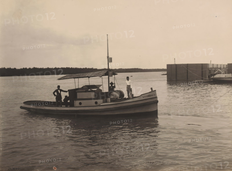 Motorboat 'Kromhout', Ocean Harbor Belawan, photograph, Motorboat 'Kromhout'. Numbered: 214. Part of the group of individual photographs regarding the construction of the harbor of Belawan on the coast off Medan, Sumatra 1921-1922., inscription: 'No. 214 Motorboat 'Kromhout'', date: '25, 9-22', Belawan, 1922-09-25, photographer, Sumatra, paper, gelatin silver print, height 160 mm x width 220 mm, harbor, building-site, steamship, motorship, economic history, overseas history, traffic and transport, infrastructure, dredging, Period, 1921 - 1922, A small wooden boat with a covered cabin sits on calm water near a larger dock structure; three men stand on the boat, two wearing hats and one holding a rope, while one man leans with a hand on the cabin roof; the boat has a rounded bow and visible deck equipment including a large barrel and a vertical mast with a small box attached; a distant treeline runs along the horizon under a mostly clear sky.