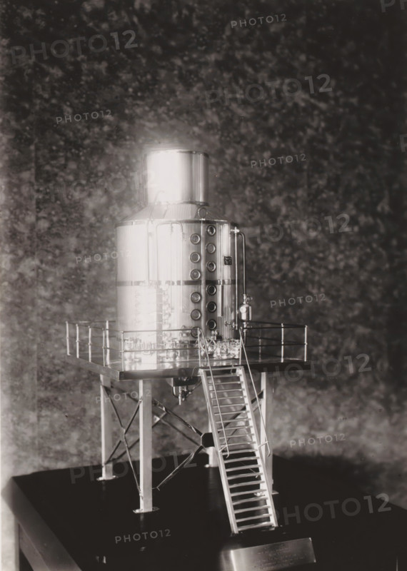Scale model of a cooking pot, photograph, without the glass cover of the display cabinet. Part of the binder with photos of machines from NV Machinefabriek Braat in Surabaya intended for the Dutch sugar industry in Java, from the period 1917-1938., Surabaya, 1917 - 1938, photographer, Java, East Java, paper, gelatin silver print, gelatin silver print on photographic paper, height 180 mm x width 130 mm, Person, NV Machinefabriek Braat, colonial system, (non-fruit) products of plants or trees: sugarcane, machines, parts of machines, tools and appliances, economic history, overseas history, sugar industry, business history, Period, A metallic cylindrical tank with a domed top sits elevated on a square platform supported by four angled legs with cross-bracing, a narrow ladder leading up to the platform, and a railing around the platform edge; the tank surface is reflective and features a vertical row of circular openings on one side and a pair of vertical pipes and valves, with various sma...