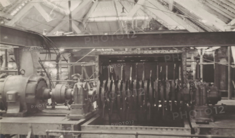 Canecutter, photograph, Electrically powered rotor of the canecutter at the Kaliwoengoe Sugar Factory west of Semarang. Part of the binder containing photographs of machines from NV Machinefabriek Braat in Surabaya intended for the Dutch sugar industry in Java, from the period 1917-1938., Java, 1917 - 1938, photographer, paper, gelatin silver print, gelatin silver print on photographic paper, height 155 mm x width 231 mm, Person, NV Machinefabriek Braat, colonial system, (non-fruit) products of plants or trees: sugarcane, factory-interior, machine shop, machines, parts of machines, tools and appliances, economic history, overseas history, sugar industry, business history, Period, A sepia-toned industrial interior showing large mechanical equipment arranged on a metal platform, including a cylindrical motor-like unit with a rounded housing on the left and a long shaft connected to a series of closely spaced disk-shaped components extending toward the right; overhead structural beams and...
