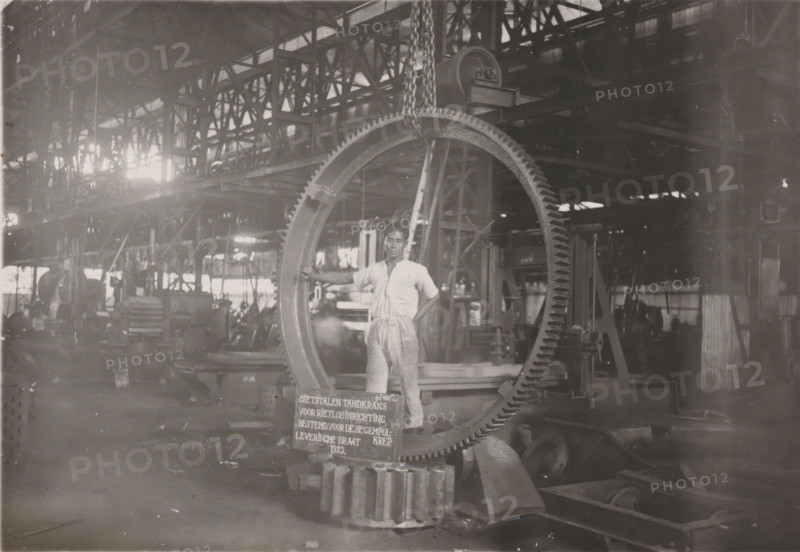 Gear ring for reed unloading device, photograph, Cast steel gear ring for a reed unloading device, manufactured in 1925 and intended for the Gempolkrep Sugar Factory. Part of the binder containing photographs of machines from NV Machinefabriek Braat in Surabaya intended for the Dutch sugar industry in Java, from the period 1917-1938., Surabaya, 1925, photographer, Java, East Java, paper, gelatin silver print, gelatin silver print on photographic paper, height 121 mm x width 173 mm, Person, NV Machinefabriek Braat, colonial system, (non-fruit) products of plants or trees: sugarcane, machines, parts of machines, tools and appliances, factory-interior, machine shop (laborers, crafts, industries, agriculture), economic history, overseas history, sugar industry, business history, Period, 1917 - 1938, A man in light-colored work clothes stands inside a large industrial workshop, his hand resting on a large circular gear that hangs from a chain; the gear has visible teeth along its outer edge...
