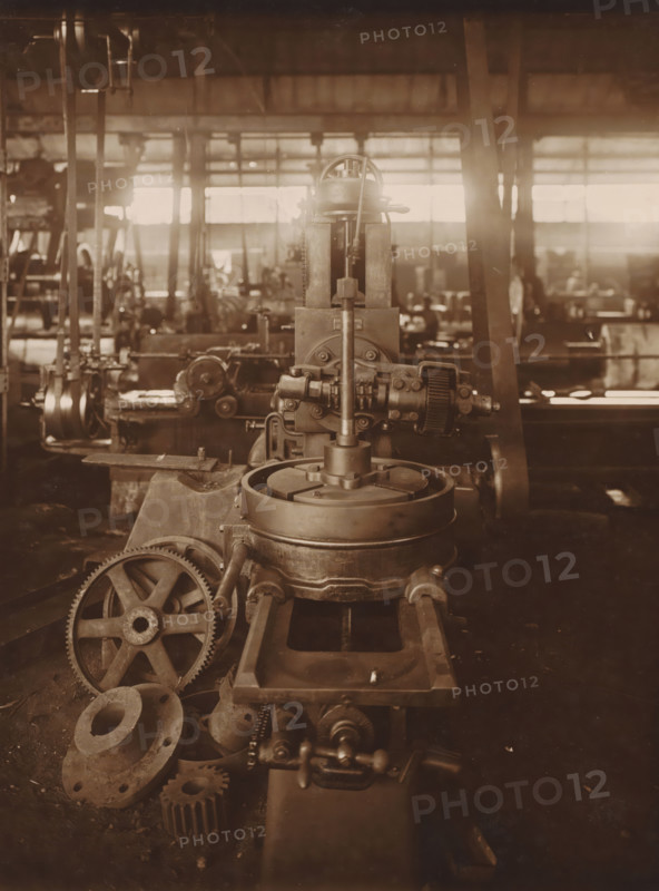 Milling machine, photograph, The first straight gear milling machine from ca. 1916. Part of the binder with photos of machines from the NV Machinefabriek Braat in Surabaya, Sukabumi, and Yogyakarta, from the period 1908-1942., Surabaya, 1919 - 1921, photographer, Java, East Java, paper, gelatin silver print, height 202 mm x width 152 mm, Person, NV Machinefabriek Braat, colonial system, factory-interior, machine shop, machines, parts of machines, tools and appliances, economic history, overseas history, business history, Period, 1908 - 1942, A sepia-toned photograph of an industrial workshop showing a large metal machine in the foreground with a vertical shaft, gears, and circular components, a set of toothed wheels and flanges resting nearby on the floor, metal rails or guides leading toward the machine, and additional blurred machines, columns, and windows in the background with light streaming through the windows.