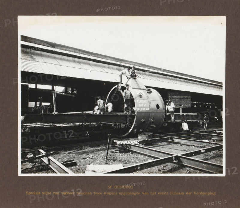 Sf. Gending, Special mode of transport suspended between two wagons of the first body of the Evaporation, album page, photograph, Album page with a photograph of the preparation for rail transport of an evaporation body. Produced by Machinefabriek Braat in Surabaya and intended for the Gending Sugar Factory. Part of the photo album Machinefabriek Braat Soerabaia-Djocja-Tegal, ca. 1916-1924., Surabaya, 1916 - 1924, photographer, Java, East Java, paper, pasteboard, gelatin silver print, developed gelatin silver print, height 162 mm x width 210 mm, height 240 mm x width 290 mm, Person, NV Machinefabriek Braat, Gending Sugar Factory, colonial system, machines, parts of machines, tools and appliances, freight train, economic history, overseas history, sugar industry, company history, Period, A black and white photograph showing a large cylindrical metal boiler-like structure mounted on a flat railcar platform with men working around and on it; the structure has riveted seams and circular op...