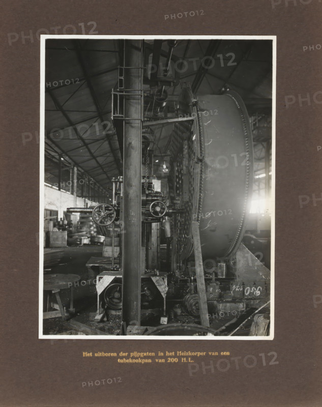 Drilling out pipe holes in the heating element of a 200 HL tubular cooking pot, album page, photograph, Album page with a photo of the operation of a machine. Produced by Machinefabriek Braat. Part of the photo album Machinefabriek Braat Soerabaia-Djocja-Tegal, ca. 1916-1924., Surabaya, 1916 - 1924, photographer, Java, East Java, paper, pasteboard, gelatin silver print, developing gelatin silver print, height 210 mm x width 162 mm, height 240 mm x width 290 mm, Person, NV Machinefabriek Braat, colonial system, factory-interior, machine shop, freight train, economic history, overseas history, sugar industry, company history, Period, A black and white photograph shows the interior of a large industrial workshop with a high pitched roof and visible metal trusses, a tall vertical metal column in the foreground with attached platforms and ladders, and a large circular metal drum with a riveted rim positioned to the right; various mechanical components, gears, pulleys, and handwheels are mou...