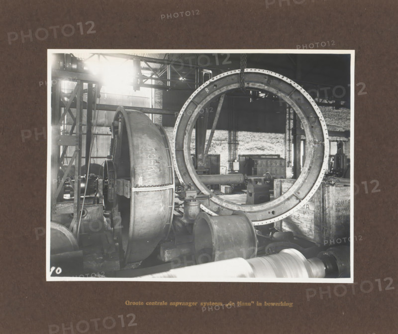 Large central juice collector system 'de Haan' in operation, album page, photograph, Album page with a photo of a machine part. Manufactured by Machinefabriek Braat. Part of the photo album Machinefabriek Braat Soerabaia-Djocja-Tegal, ca. 1916-1924., Surabaya, 1916 - 1924, photographer, Java, East Java, paper, pasteboard, gelatin silver print, developing gelatin silver print, height 157 mm x width 207 mm, height 240 mm x width 290 mm, Person, NV Machinefabriek Braat, colonial system, factory-interior, machine shop, machines, parts of machines, tools and appliances, economic history, overseas history, sugar industry, company history, Period, A black and white photograph shows an industrial workshop interior with large metal machinery and components. A large circular metal ring with bolt holes dominates the right side, suspended from a chain attached above. To the left of the ring is a curved metal housing and visible gears and shafts. Various cylindrical metal parts and pipes lie on the...