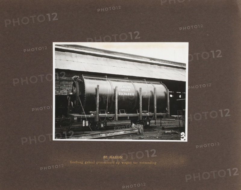 Sf. Maron Cooling Trough fully mounted on wagon for shipment, album page, photograph, Album page with a photo of the transport of a machine part. Manufactured by Machinefabriek Braat and intended for the Maron Sugar Factory. Part of the photo album Machinefabriek Braat Soerabaia-Djocja-Tegal, ca. 1916-1924., Surabaya, 1916 - 1924, photographer, Java, East Java, paper, pasteboard, gelatin silver print, developing gelatin silver print, height 115 mm x width 157 mm, height 240 mm x width 290 mm, Person, NV Machinefabriek Braat, Maron Sugar Factory, colonial system, machines, parts of machines, tools and appliances, freight train, economic history, overseas history, sugar industry, company history, Period, A black and white photograph mounted on a dark board shows a large cylindrical metal tank mounted horizontally on a rail wagon inside an industrial shed with a corrugated roof, the tank is supported by vertical wooden or metal braces and secured with chains, one end of the tank features ...