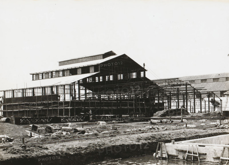 Marine Establishment, Surabaya, Submarine Repair Workshop, album page, photograph, Album page with a photo of the skeleton of a Navy submarine workshop in Surabaya, under construction. Part of the photo album Machinefabriek Braat Soerabaia-Djocja-Tegal, ca. 1916-1924., 1916 - 1924, photographer, Java, East Java, paper, pasteboard, gelatin silver print, developed gelatin silver print, height 179 mm x width 230 mm, height 240 mm x width 290 mm, Person, NV Machinefabriek Braat, Royal Netherlands Navy, navy, making the carcass (, building activities), colonial system, submarine, economic history, overseas history, architecture, business history, Period, A black-and-white photograph shows a large industrial building under construction with exposed steel framework and scaffolding, a multi-tiered roof structure with windows and open sections, and temporary supports and beams surrounding the structure; in the foreground there is a muddy construction site with scattered barrels, wooden planks, ...