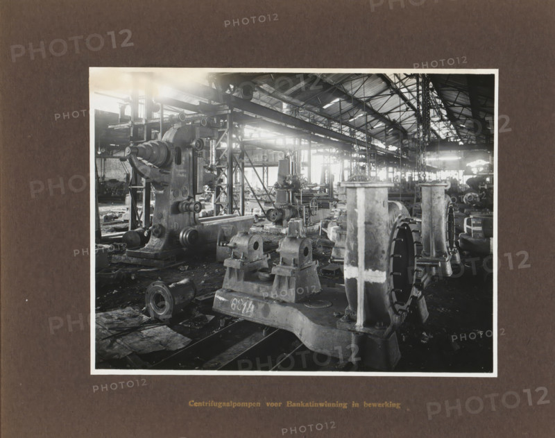 Centrifugal pumps for Banka tin extraction in progress, album page, photograph, Album page with a photo of the manufacture of pumps. Manufactured by Machinefabriek Braat and intended for Banka tin extraction. Part of the photo album Machinefabriek Braat Soerabaia-Djocja-Tegal, ca. 1916-1924., Surabaya, 1916 - 1924, photographer, Java, East Java, paper, pasteboard, gelatin silver print, developed gelatin silver print, height 157 mm x width 208 mm, height 240 mm x width 290 mm, Person, Banka tin mining, NV Machinefabriek Braat, colonial system, factory-interior, machine shop, machines, parts of machines, tools and appliances, economic history, overseas history, sugar industry, company history, Period, A black and white photograph shows an industrial workshop filled with large cast metal machinery and heavy components arranged on the floor and on platforms, with a prominent machine casting in the foreground featuring a rounded housing and cylindrical protrusions, flanged circular parts, a...
