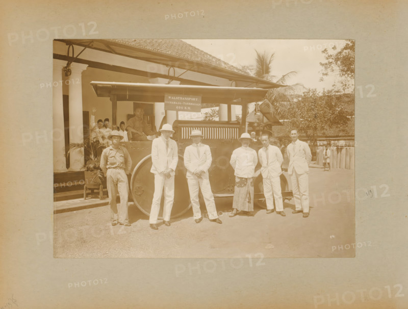 Roller transport, album page, photograph, Album page with a photo of a roller transported from Surabaya to Tasikmalaya in West Java. Dutch and Indonesian men are posing by the roller; this is likely in Tasikmalaya. Part of a photo album with photos of machines and constructions of the NV Machinefabriek Braat in Surabaya, Sukabumi, and Tegal from the period 1931-1937., Java, 1931 - 1937, photographer, paper, pasteboard, daylight gelatin silver print, height 167 mm x width 232 mm, height 250 mm x width 320 mm, Person, NV Machinefabriek Braat, other motor-vehicles not meant or mentioned above, economic history, overseas history, business history, Period, A sepia photograph showing a group of people standing in front of a large vehicle with a visible wheel and a canopy, with a building featuring columns and a tiled roof behind them; several men in light-colored suits and hats stand in a row at the front, alongside a man in a darker work shirt and a man wearing a patterned skirt and light j...