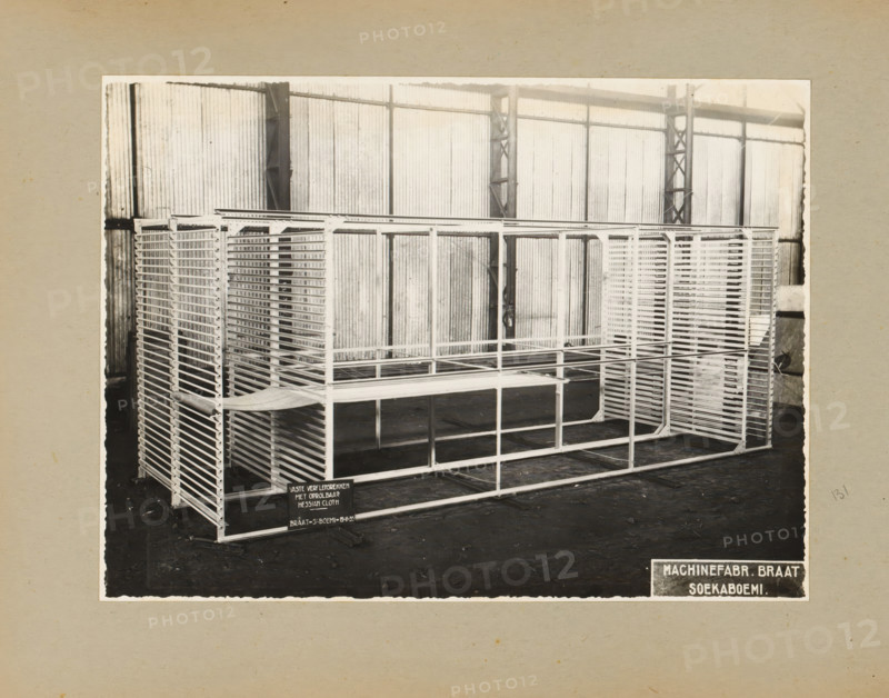 Paint lamination racks, album page, photograph, Album page with a photo of 'fixed paint lamination racks with rollable Hessian Cloth', November 19, 1935. Part of a photo album with photos of machines and constructions of the NV Machinefabriek Braat in Surabaya, Sukabumi and Tegal from the period 1931-1937., Sukabumi, 1935-11-19, photographer, Java, West-Java, paper, pasteboard, gelatin silver print, developing gelatin silver print, height 168 mm x width 227 mm, height 250 mm x width 320 mm, Person, NV Machinefabriek Braat, factory-interior, machine shop, machines, parts of machines, tools and appliances, economic history, overseas history, company history, Period, 1931 - 1937, A black and white photo shows a large metal shelving structure with multiple horizontal slats and rectangular frames arranged in rows, placed on a dark floor inside a corrugated metal-walled interior with vertical support beams and truss-like columns; the shelving has open grid sides and stacked tray-like levels,...