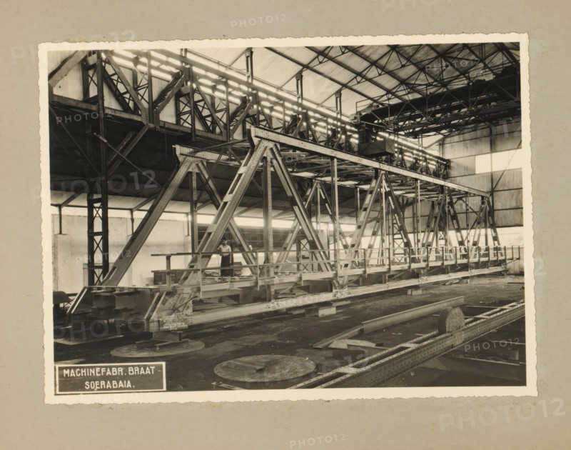 Railway bridge, album page, photograph, Album page with a photo of a truss bridge (railway bridge with footbridge) intended for the State Railways, January 29, 1937. Part of a photo album with photos of machines and constructions of the NV Machinefabriek Braat in Surabaya, Sukabumi, and Tegal from the period 1931-1937., Surabaya, 1937-01-29, photographer, Java, East Java, paper, pasteboard, gelatin silver print, developing gelatin silver print, height 168 mm x width 228 mm, height 250 mm x width 320 mm, Person, Company for the Exploitation of State Railways, NV Machinefabriek Braat, railway, train, factory-interior, machine shop, economic history, overseas history, bridge construction, company history, Period, 1931 - 1937, A sepia-toned photograph of an industrial interior showing a large metal truss bridge structure supported by triangular steel frames and riveted beams inside a high-roofed building with exposed roof trusses and corrugated wall panels; walkways and railings run along ...