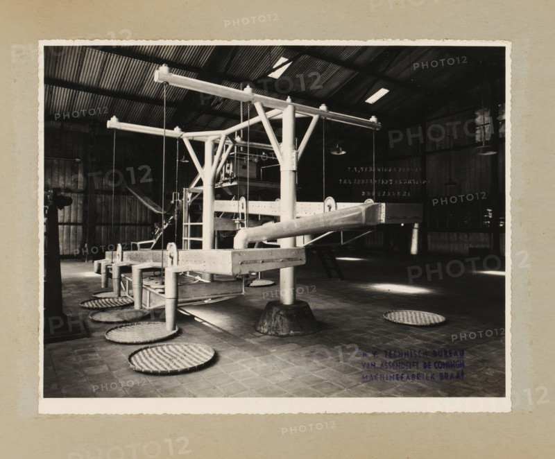 Installation, album page, photograph, Album page with a photo of an installation with conveyor belts. Designed for or by NV Technisch Bureau Van Assendelft De Coningh. Part of a photo album with photos of machines and structures of the NV Machinefabriek Braat in Surabaya, Sukabumi, and Tegal from the period 1931-1937., name: 'NV Technisch Bureau Van Assendelft De Coningh Sukabumi', name: 'NV Technisch Bureau Van Assendelft De Coningh Machinefabriek Braat', 1931 - 1937, photographer, Java, West-Java, paper, pasteboard, gelatin silver print, height 170 mm x width 216 mm, height 250 mm x width 320 mm, Person, NV Machinefabriek Braat, factory-interior, machine shop, machines, parts of machines, tools and appliances, economic history, overseas history, company history, Period, A black and white photograph of an industrial interior showing a large metal framework with horizontal beams and vertical supports, pipes and rods connected to a central column, suspended lights hanging from the ceili...