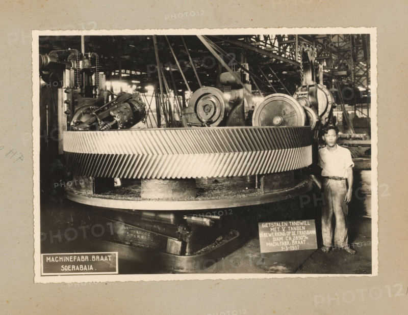 Gear, album page, photograph, Album page with a photo of a cast steel gear being worked on the milling machine in 1937 and intended for the Gempolkerep Sugar Factory. Part of a photo album with photos of machines and constructions of the NV Machinefabriek Braat in Surabaya, Sukabumi, and Tegal from the period 1931-1937., Surabaya, 1937-03-01, photographer, Java, East Java, paper, pasteboard, gelatin silver print, developing gelatin silver print, height 168 mm x width 227 mm, height 250 mm x width 320 mm, Person, NV Machinefabriek Braat, parts of machines, economic history, overseas history, sugar industry, company history, Period, 1931 - 1937, A black-and-white photograph showing a large industrial gear with diagonal toothed ridges mounted on a circular base, surrounded by machinery components including pulleys, chains, gears, and structural beams; a man wearing a light-colored short-sleeved shirt and trousers stands beside the gear near the right edge of the image; the background reve...