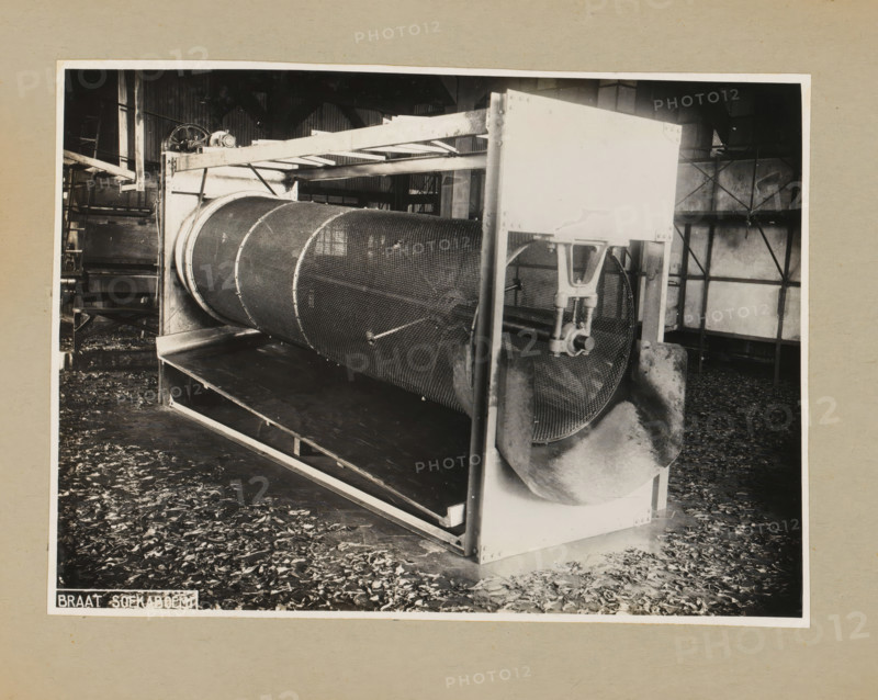 Drum, album page, photograph, Album page with a photo of a drum for a machine. Part of a photo album with photos of machines and constructions of the NV Machinefabriek Braat in Surabaya, Sukabumi, and Tegal from the period 1931-1937., 1931 - 1937, photographer, Java, West-Java, paper, pasteboard, gelatin silver print, developing gelatin silver print, height 169 mm x width 232 mm, height 250 mm x width 320 mm, Person, NV Machinefabriek Braat, factory-interior, machine shop, machines, parts of machines, tools and appliances, economic history, overseas history, company history, Period, A black and white photograph of an industrial interior showing a large cylindrical metal drum with a mesh surface mounted horizontally within a rectangular metal frame, supported by vertical posts and brackets, with a curved metal chute attached to one end of the drum and a clamping mechanism visible nearby; the drum sits above a flat tray or conveyor and leaves scattered across the floor; background elemen...