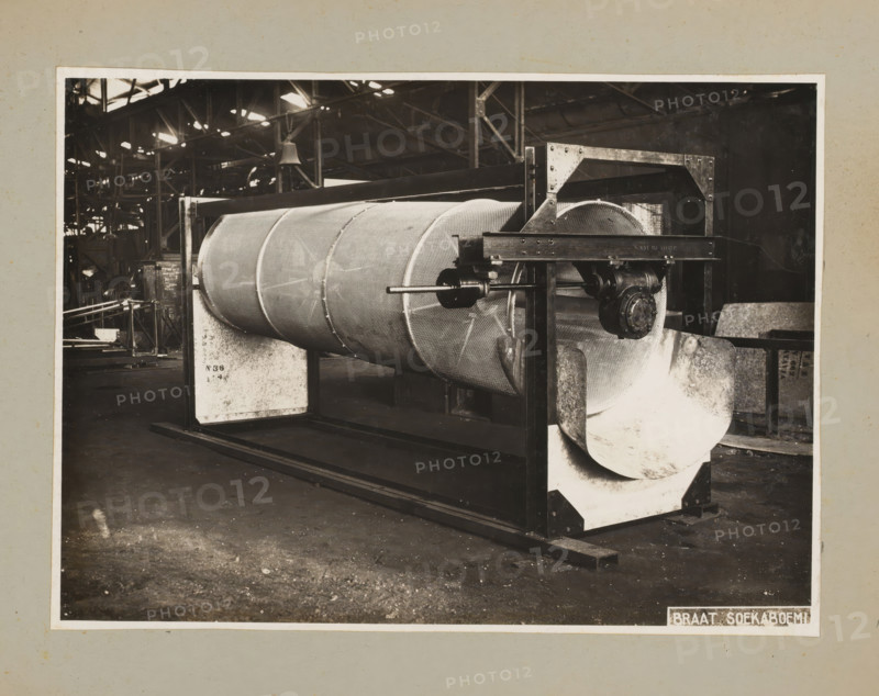 Drum, album page, photograph, Album page with a photo of a drum for a machine. Part of a photo album with photos of machines and constructions of the NV Machinefabriek Braat in Surabaya, Sukabumi, and Tegal from the period 1931-1937., 1931 - 1937, photographer, Java, West-Java, paper, pasteboard, gelatin silver print, developed gelatin silver print, height 172 mm x width 236 mm, height 250 mm x width 320 mm, Person, NV Machinefabriek Braat, factory-interior, machine shop, machines, parts of machines, tools and appliances, economic history, overseas history, company history, Period, A large cylindrical metal drum with a textured surface is mounted horizontally within a rectangular metal frame, visible from an angled front view inside a spacious industrial building. The drum shows segmented reinforcement bands and is supported by padded cradle blocks at each end, with a central shaft and bearing assembly projecting through the frame. The frame includes vertical and horizontal beams with ...