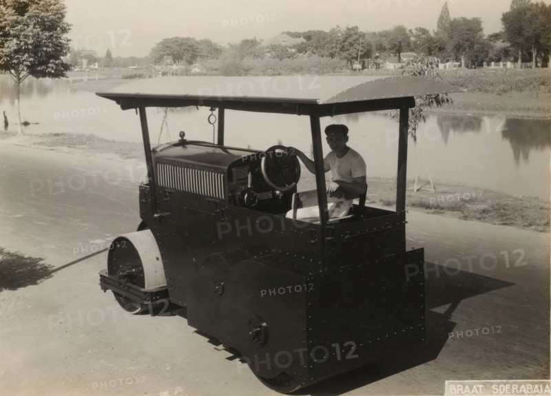 Roller with operator, album page, photograph, Album page with a photo of a roller with the Javanese operator. Part of a photo album with photos of machines and constructions of the NV Machinefabriek Braat in Surabaya, Sukabumi, and Tegal from the period 1931-1937., Surabaya, 1931 - 1937, photographer, Java, East Java, paper, pasteboard, gelatin silver print, height 166 mm x width 229 mm, height 250 mm x width 320 mm, Person, NV Machinefabriek Braat, machine driven by steam, other motor-vehicles not meant or mentioned above, economic history, overseas history, business history, Period, 1931 - 1938, A vintage road roller with a flat canopy and riveted metal panels is parked on a paved surface next to a body of water, a man in a light shirt sits at the controls holding the steering wheel, trees and low vegetation line the far bank with reflections visible in the calm water, and a single leafy tree stands near the water’s edge on the left side of the scene.