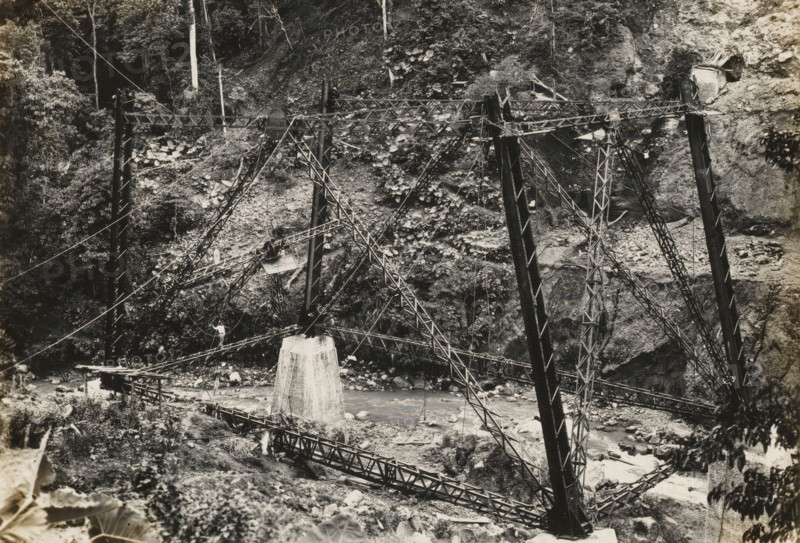 Railway bridge over the Ci Somang under construction, album page, photograph, Album page with a photo of the railway bridge over the Ci Somang in Preanger under construction in 1931-1932. The three piers were supplied in 1931 by Machinefabriek Braat. Part of a photo album with photos of machines and structures of the NV Machinefabriek Braat in Surabaya, Sukabumi, and Tegal from the period 1931-1937., date: 'Tji Somang 26-11-31', Preanger, 1931-11-26, photographer, Java, West-Java, paper, pasteboard, gelatin silver print, developed gelatin silver print, height 178 mm x width 243 mm, height 250 mm x width 320 mm, Other copies of the photos of the construction of this bridge in album NG-2002-84-C., Person, NV Machinefabriek Braat, railway, tramway, rack railway, bridge, economic history, overseas history, bridge building, company history, Ci Somang, Period, 1931 - 1937, A black and white photograph mounted on a beige matte shows a partially constructed metal bridge spanning a rocky river ...