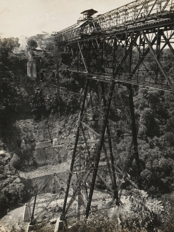 Railway bridge over the Ci Somang under construction, album page, photograph, Album page with a photo of the railway bridge over the Ci Somang in Preanger under construction in 1931-1932. The three piers were supplied in 1931 by Machinefabriek Braat. Part of a photo album with photos of machines and structures of the NV Machinefabriek Braat in Surabaya, Sukabumi, and Tegal from the period 1931-1937., date: 'Tji Somang 26-11-'31', Preanger, 1931-11-16, photographer, Java, West-Java, paper, pasteboard, gelatin silver print, developed gelatin silver print, height 240 mm x width 179 mm, height 250 mm x width 320 mm, Other copies of the photos of the construction of this bridge in album NG-2002-84-C., Person, NV Machinefabriek Braat, railway, tramway, rack railway, bridge, economic history, overseas history, bridge building, company history, Ci Somang, Period, 1931 - 1937, A black-and-white photograph showing a large metal truss bridge spanning a deep rocky gorge, with tall lattice steel su...